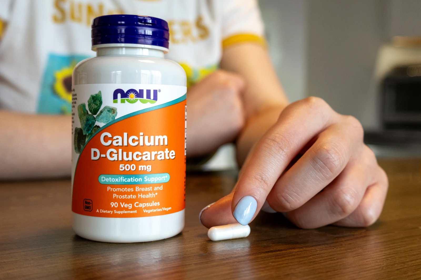 A bottle of NOW Calcium D-Glucarate with a single capsule on a wood table